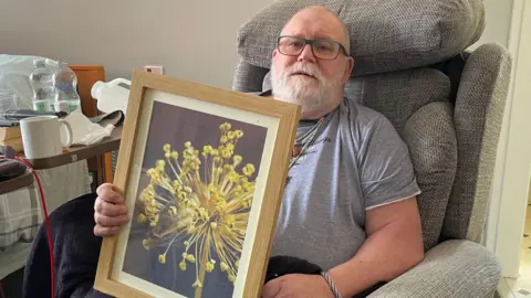 Tom MacDougall/BBC Peter Hardy holding up one of his photographs, which captured a mouse hiding inside a flower.