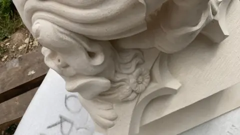BBC/Jack Hadaway-Weller An intricately carved stone corbel features a sculpted face surrounded by elegant scrollwork and floral motifs. The craftsmanship highlights fine details, including flowing curves, textured surfaces, and decorative architectural elements.
