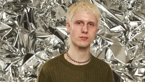 Jake Moran looks at the camera. He has short, bleach blonde hair. He is wearing a knitted green jumper and metallic necklace and silver hoop earrings. 