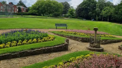 Wellingborough Town Council A garden, with several flower beds, all in bloom, a green bench, and a path that looks like its made from lots of slabs. There are building in the distance, to the left and trees around the area. There are large areas of green grass. An ornament is slightly to the right, with a small fence round it. 