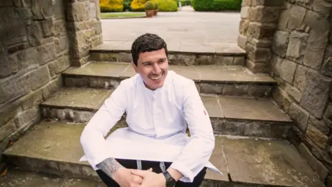 A man with dark hair, wearing a white chef's coat, sits on a flight of stone steps. A garden is visible behind the steps.