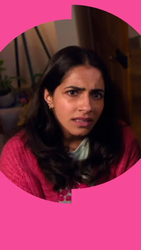 A woman with long, dark hair wearing a bright pink jumper looks confused