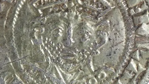 A close-up of the front a 13th Century silver gilt coin brooch showing the face of a bearded French bishop