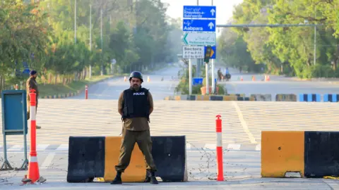 Reuters A man wearing uniform which says the word POLICE on it stands in front of a cordoned-off road.