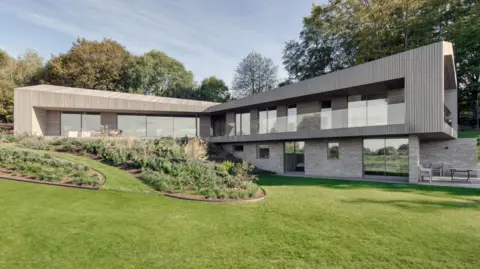 RIBA A split-level modernist house built into a bank with two visible wings at a right angle to each other with a garden and grass in the foreground 