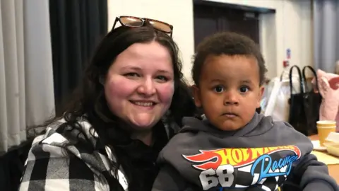 A woman in her mid-20s with shoulder-length dark hair and sunglasses on top of her head is smiling at the camera. She is wearing a white and black checked top. On her lap is her toddler son who is wearing a grey Hot Wheels Racing hoodie.