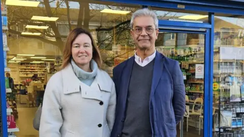 Marie Goldman and Dipak Pau wearing coats outside a pharmacy in Essex. They are both smiling at the camera