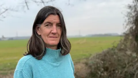 Steve Hubbard/BBC Rosie Pearson standing by a field with pylons in the background. She has shoulder-length brown hair and is wearing a light blue jumper.