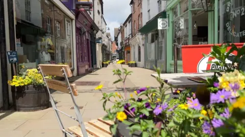 BBC An empty street with shops either side of the path. In the foreground are plants and a table and chairs. Between the shops are beige slabs of paving.