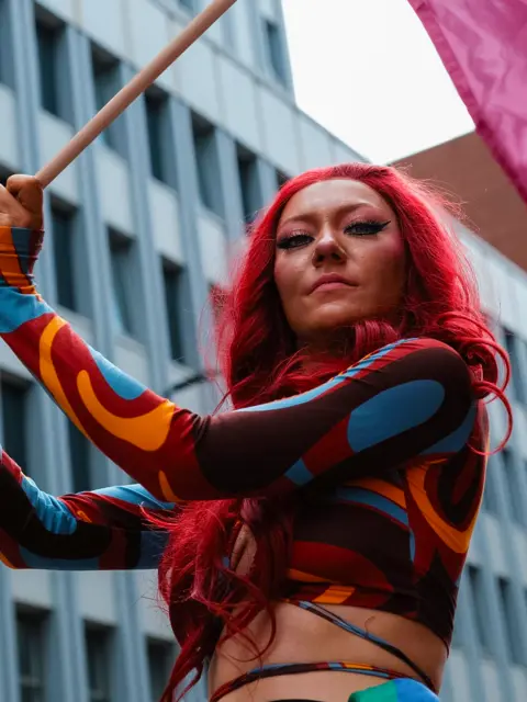 A parade participant with long bright red hair holds a pink flag aloft while wearing a patterned outfit featuring swirling blue, orange and brown designs.