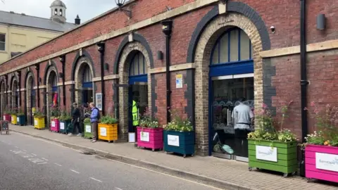 BBC Pannier Market in north Devon - a long one story brown brick building with different coloured potted plant holders, seven entries, grey skies