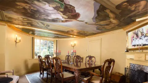 Savills A dining room with a ceiling painting of classical gods. On the right hand side of the room above the fire place is a framed painting of Alice in Wonderland at the Mad Hatter's Tea Party.