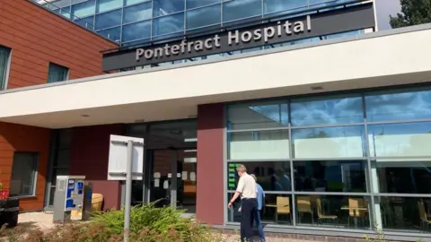 Two people walk into the entrance of a large modern-looking building. A sign out the front reads 'Pontefract Hospital'. 