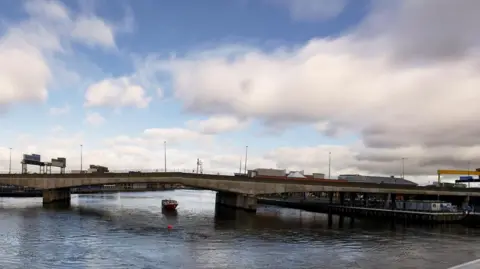 Getty Images A wide shot of the Lagan Bridge in Belfast. It's a large concrete bridge over the Lagan River. In the distance is one of the yellow Harland and Wolff cranes, and the top of the SSE Arena and Odyssey pavillion buildings. It is a cloudy day, with some breaks of blue sky.