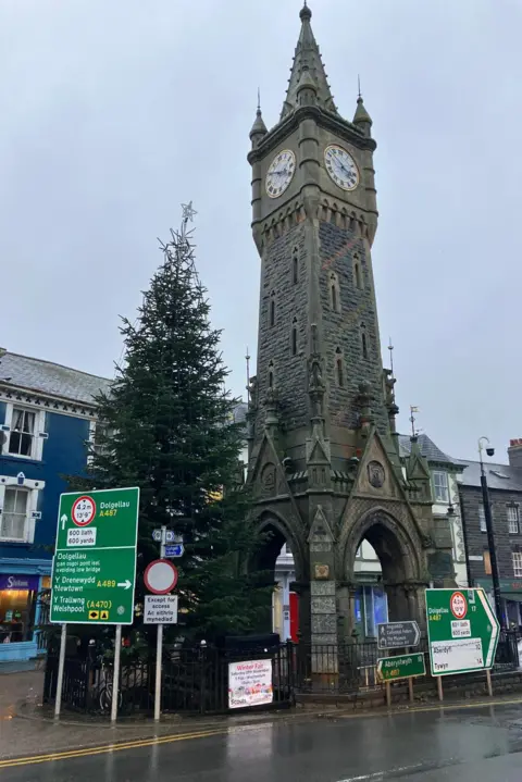 The mayor's tree in the middle of the high street. It sits beside a clock tower and in front of a green road directions sign. 