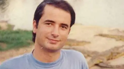 Acun Medya A colour photograph showing a young adult sitting outdoors near a body of water, wearing a light blue T-shirt. Several crocodiles or alligator-like reptiles are visible on the bank behind.