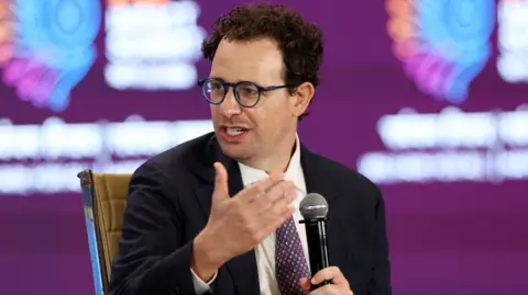 Dario Amodei, Anthropic's CEO, speaking on stake while holding a microphone, wearing a black suit jacket, white shirt and red and navy spotted neck tie.