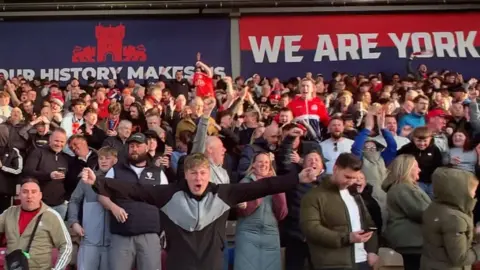 A group of mixed gender football fans celebrate a victory in the stands of a stadium. Behind the group, two large signs in red and blue read: We Are York and Our History Makers.