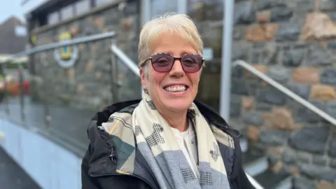 Deputy Rhona Humphreys - A woman smilking with bared teeth, with short blonde hair, wearing red tinted rimmed glasses, with multi-colours on the top rim. She has a cream and blue scarf on, with a blue coat and a white blouse