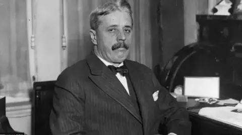 Getty Images A black and white picture from the early 20th Century of a man sitting on a chair in an office. He is wearing a pin-striped suit and a bow tie. He is looking into the camera but is not smiling. 