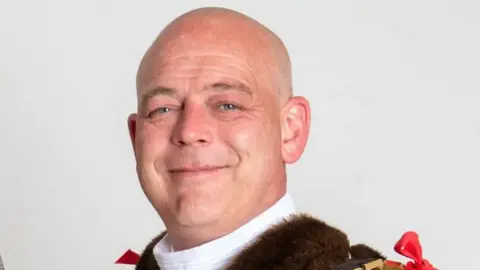 City of Wolverhampton Council A headshot of Greg Brackenridge, wearing mayoral clothes. He has a bald head and slight smile at the camera.