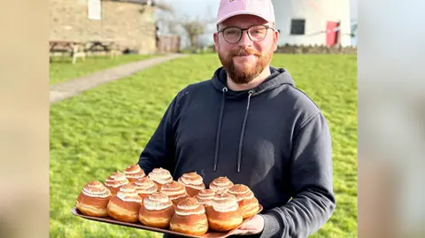 Richard Holt Richard Holt wearing a pink cap hat, a dark hoodie and glasses. He is holding a tray of doughnuts with cream topping and chocolate dusting. 