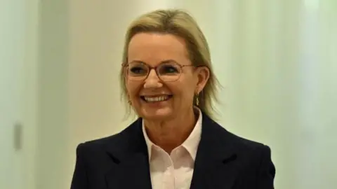 Reuters Liberal leader Sussan Ley, wearing a white collared shirt and black blazer, walking down corridor, smiling and looking off camera to the left