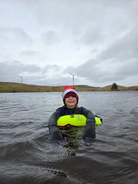 Fiona Baird Young girl wearing a Santa hat, holding onto an inflatable lift raft, smiling while floating in a loch.