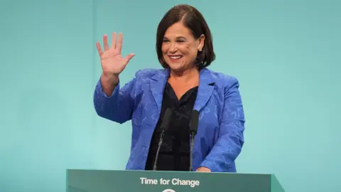 Mary Lou McDonald, President of PA Media Sinn Fein, standing on a light green podium, delivering her speech to Sinn Fein Ard Fheis. She wears a blue patterned suit jacket and a black top. 