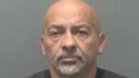 A mugshot of John Aneli who is bald with a grey goatee. 