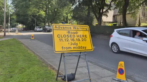 A yellow traffic sign next to road leading to a park gate, saying the route will closes on concert days