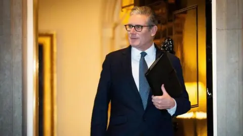 PA Media Keir Starmer walks out of the door to Number 10 Downing Street. He has short grey hair, glasses, and is wearing a navy suit and blue tie.