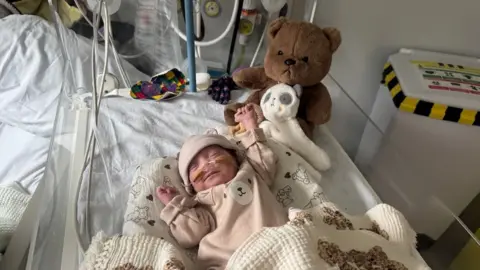 A premature baby wearing a light brown sleepsuit with a bear face on his tummy, sleeps in his hospital cot. He has a cream blanket with teddy bears over his legs. He shares the cot with two cuddly toys.