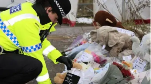 GMP A GMP officer kneels down to place a card alongside other tributes to the baby, which include flowers and stuffed animals on a cold day, with frost and a forensic tent seen in the background. 