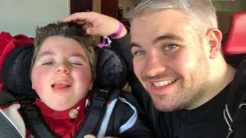 Paul Woodward Paul Woodward is sitting next to his son Kalel with his hand resting on his head. Kalel is sticking his tongue out while Paul is smiling. 