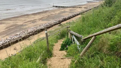 Owen Sennitt/BBC A set of handrails badly damaged can be seen next to a series of steps that lead down to a sandy beach in Mundesley, with sea defences and groynes visible in the background