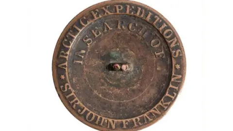 Bearnes, Hampton and Littlewood The button. It is brown. It says 1852 on it and has writing going along various bits of it. It is on a white background.