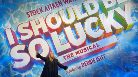 BBC/Chloee French Debbie Isitt standing in front of the title curtain of her musical I Should Be So Lucky.
