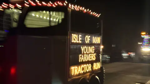 A trailer in the convoy with Isle of Man You Farmers Tractor Run in lights on the side.