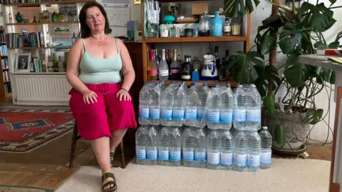 A woman is sitting on a wooden chair in her kitchen. She is wearing a light green tank top and bright pink shorts. Next to her are rows of water bottles stacked on top of each other. In the background there are bookshelves with some books, plants, and other household items. There are also several bottles and containers on the shelves as well as a large green house plant.