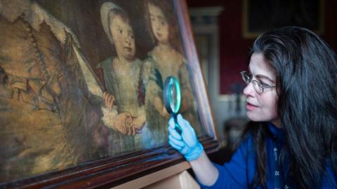 A curator uses a magnifying glass to inspect an 18th Century print of a portrait of Charles I's three eldest children 