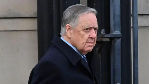 Pacemaker William Lloyd-Lavery, a man with grey hair, a dark jacket, blue shirt and blue tie, pictured beside a grey brick building.