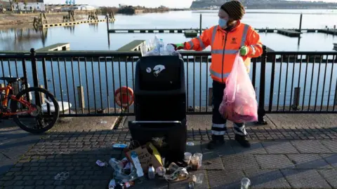 Getty Images A refuse worker in orange hi-vis holds a clear plastic binbag and empties a bin at Cardiff Bay. The water is behind him.
