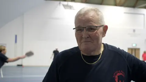 Jamie Niblock/BBC Steve has white hair and is wearing glasses. He is wearing the team's alternative training kit. It is navy blue with a pink logo. 