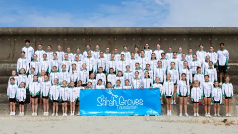 The Sarah Groves Foundation A group of young dancers standing with a Sarah Groves Foundation banner. 