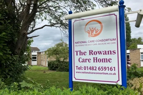 BBC / David Rimmington Brick one-storey building with windows and surrounded by trees. There is a sign in front which reads National Care Consortium, delivering the best care because we care, The Rowans Care Home. Tel: 01482 659161. www.nationalcareconsortium.co.uk.