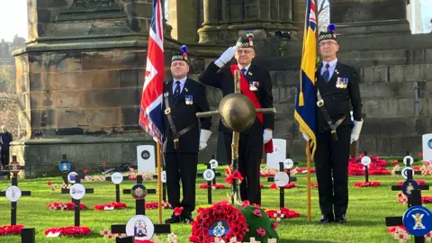 PA Media Three uniformed men stand in a Remembrance garden in Edinburgh Princes Street flanked by two flags, one of which is a Union Flag. They are all saluting and there are small crosses on the ground all around them. 