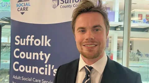 Vikki Irwin/BBC A man standing in a room next to a sign which reads "Suffolk County Council's Adult Social Care services". He is wearing a blue jacket, a white shirt and a blue and pink tie.