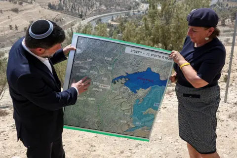 Reuters A man in a suit holds a large map along with a woman. They are stood on a dusty barren hill. 