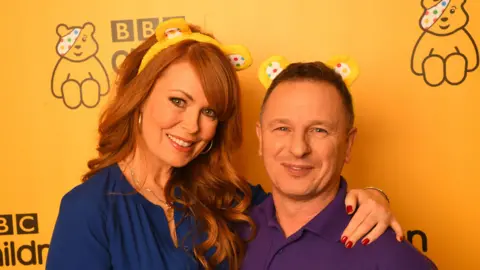 Gabrielle Blackman (L) and Mark Millar of DIY SOS backstage at BBC Children in Need's 2019 Appeal night at Elstree Studios. Mark is wearing a purple shirt and Gabrille is wearing a navy dress, they both have Pudsey's ear headband on and are staying in front of Pudsey bear and BBC Children in Need logo on a yellow wall.